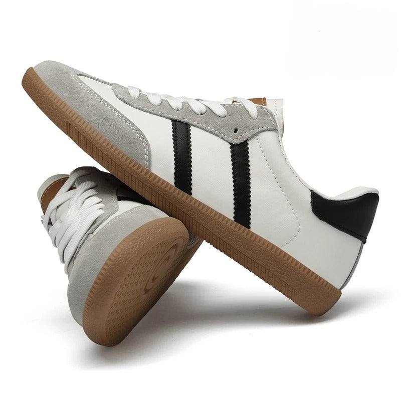 A pair of low-top sneakers shown on a white background, with one shoe standing upright and the other tilted underneath. The shoes are primarily white with light gray suede overlays, black side stripes, a black heel tab, and a brown gum sole.